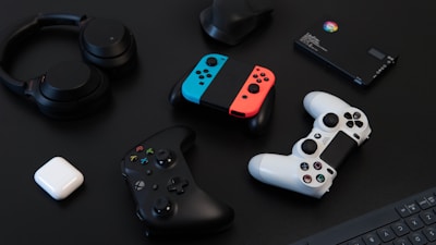 A collection of gaming devices and accessories on a dark background, including headphones, a gaming controller, and a Nintendo Switch with colored joy-cons. A white earbud case and a small electronic device are also present.