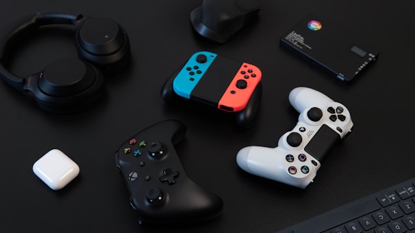 A collection of gaming devices and accessories on a dark background, including headphones, a gaming controller, and a Nintendo Switch with colored joy-cons. A white earbud case and a small electronic device are also present.
