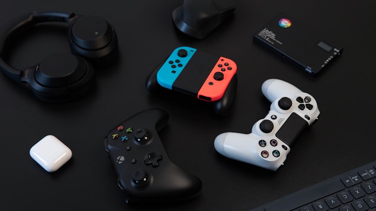 A collection of gaming devices and accessories on a dark background, including headphones, a gaming controller, and a Nintendo Switch with colored joy-cons. A white earbud case and a small electronic device are also present.