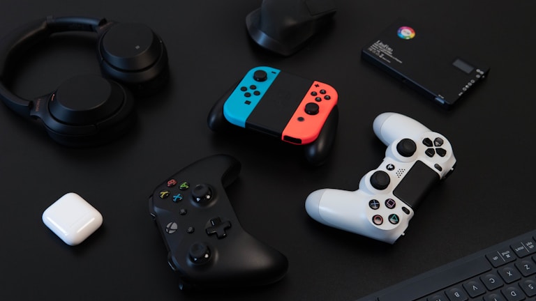 A collection of gaming devices and accessories on a dark background, including headphones, a gaming controller, and a Nintendo Switch with colored joy-cons. A white earbud case and a small electronic device are also present.