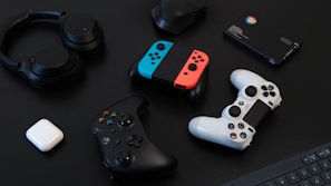A collection of gaming devices and accessories on a dark background, including headphones, a gaming controller, and a Nintendo Switch with colored joy-cons. A white earbud case and a small electronic device are also present.