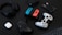 A collection of gaming devices and accessories on a dark background, including headphones, a gaming controller, and a Nintendo Switch with colored joy-cons. A white earbud case and a small electronic device are also present.