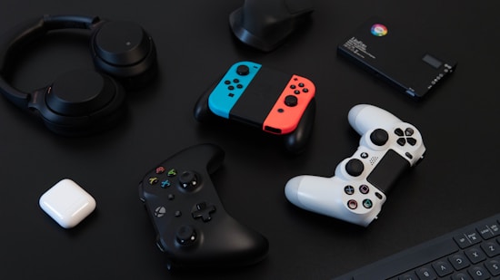 A collection of gaming devices and accessories on a dark background, including headphones, a gaming controller, and a Nintendo Switch with colored joy-cons. A white earbud case and a small electronic device are also present.