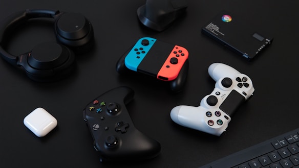 A collection of gaming devices and accessories on a dark background, including headphones, a gaming controller, and a Nintendo Switch with colored joy-cons. A white earbud case and a small electronic device are also present.