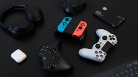 A collection of gaming devices and accessories on a dark background, including headphones, a gaming controller, and a Nintendo Switch with colored joy-cons. A white earbud case and a small electronic device are also present.