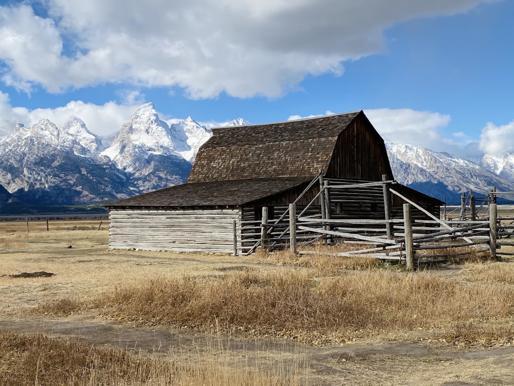 brown wooden barn on brown grass field near snow covered mountain under blue and white cloudy