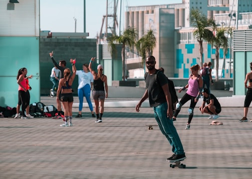 A group of people are gathered in an outdoor urban setting, some are talking and interacting while others are engaged in various activities like skateboarding and exercising. The scene is lively with a mix of people using athletic wear, backpacks are placed on the ground. There are palm trees and modern architectural buildings in the background.