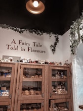 A cozy display inside a shop features wooden shelves filled with various baked goods and jars. The shelves are labeled 'The Vegan Corner' and are accompanied by a sign that reads 'A Fairy Tale Told in Flavours.' Green vines decorate the walls and ceiling, and a warm light hangs overhead, creating an inviting atmosphere.