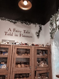 A cozy display inside a shop features wooden shelves filled with various baked goods and jars. The shelves are labeled 'The Vegan Corner' and are accompanied by a sign that reads 'A Fairy Tale Told in Flavours.' Green vines decorate the walls and ceiling, and a warm light hangs overhead, creating an inviting atmosphere.