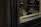 A person, dressed in a suit, is sitting indoors reading a document. The scene is dimly lit with reflections on glass, and the background shows a set of doors leading outside.