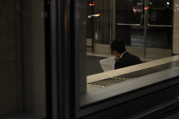 A person, dressed in a suit, is sitting indoors reading a document. The scene is dimly lit with reflections on glass, and the background shows a set of doors leading outside.