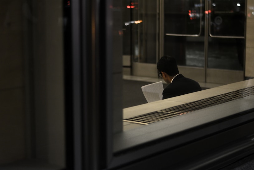 A person, dressed in a suit, is sitting indoors reading a document. The scene is dimly lit with reflections on glass, and the background shows a set of doors leading outside.