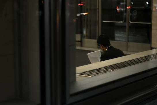 A person, dressed in a suit, is sitting indoors reading a document. The scene is dimly lit with reflections on glass, and the background shows a set of doors leading outside.
