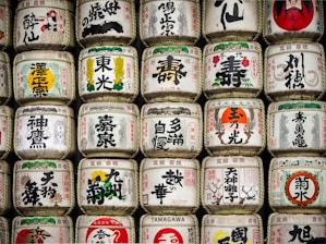 Rows of traditional Japanese sake barrels, decorated with intricate kanji and colorful symbols, are stacked neatly. Each barrel features unique designs and calligraphy, representing different breweries or styles of sake.