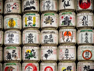 Rows of traditional Japanese sake barrels, decorated with intricate kanji and colorful symbols, are stacked neatly. Each barrel features unique designs and calligraphy, representing different breweries or styles of sake.