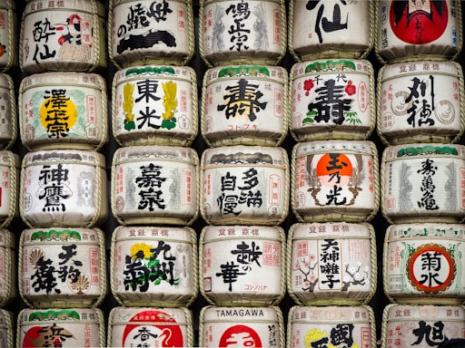 Rows of traditional Japanese sake barrels, decorated with intricate kanji and colorful symbols, are stacked neatly. Each barrel features unique designs and calligraphy, representing different breweries or styles of sake.