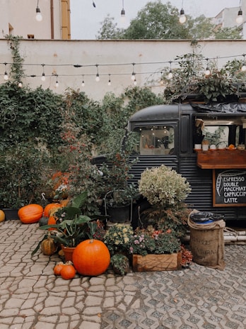 A quaint outdoor setting featuring a vintage food truck surrounded by lush greenery and a variety of pumpkins and plants. String lights hang above, adding a warm, inviting ambiance. A chalkboard menu displays coffee options, and a burlap sack adds a rustic touch to the decor.