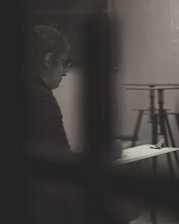 A person is sitting alone, appearing to write or sketch on a clipboard. The image is in black and white, with soft lighting creating a contemplative and introspective atmosphere. The background includes blurred elements, like a stand, which enhance the focus on the individual.