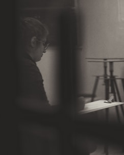 A person is sitting alone, appearing to write or sketch on a clipboard. The image is in black and white, with soft lighting creating a contemplative and introspective atmosphere. The background includes blurred elements, like a stand, which enhance the focus on the individual.