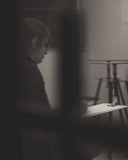 A person is sitting alone, appearing to write or sketch on a clipboard. The image is in black and white, with soft lighting creating a contemplative and introspective atmosphere. The background includes blurred elements, like a stand, which enhance the focus on the individual.
