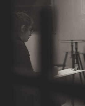 A person is sitting alone, appearing to write or sketch on a clipboard. The image is in black and white, with soft lighting creating a contemplative and introspective atmosphere. The background includes blurred elements, like a stand, which enhance the focus on the individual.