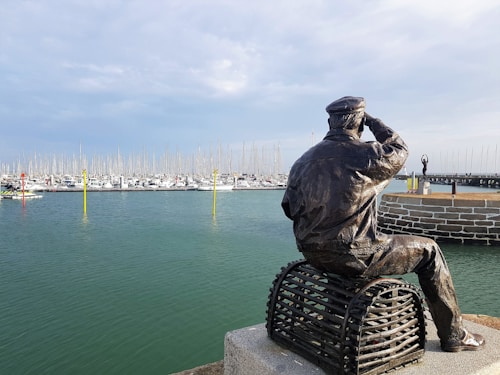 A bronze statue of a sailor sits on a stack of wire traps, overlooking a harbor filled with numerous sailboats. The sailor has one hand raised to his eyes as if searching the horizon. The water is a calm turquoise color, and a line of masts creates a rhythmic pattern against the cloudy sky.