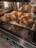 Five golden-brown croissants are baking in an oven, placed on a baking tray lined with parchment paper. The oven has a sleek, digital control panel with several icons and a timer displaying '00:05'. The interior is well-lit, highlighting the texture of the pastries.
