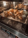 Five golden-brown croissants are baking in an oven, placed on a baking tray lined with parchment paper. The oven has a sleek, digital control panel with several icons and a timer displaying '00:05'. The interior is well-lit, highlighting the texture of the pastries.