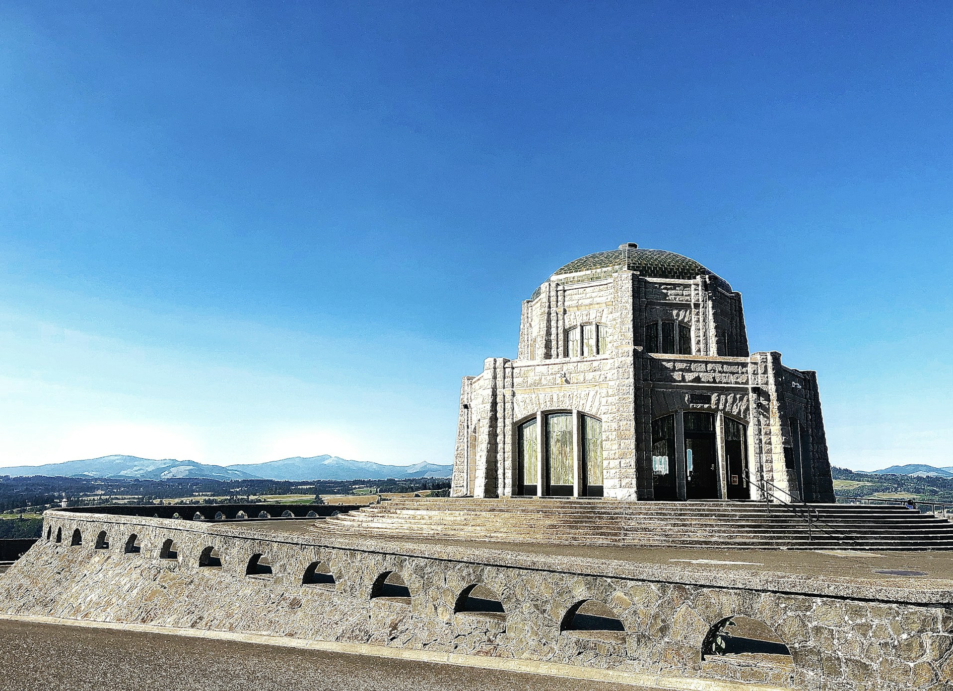 A large stone observatory-like structure with an octagonal shape and multiple arched windows, situated on a stone platform with a clear blue sky and distant mountain range in the background.