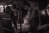 A person is seated inside a vehicle, likely a tuk-tuk or auto rickshaw. The individual appears to be in a pensive or contemplative mood, with a visible 'For Hire' sign on the vehicle. The setting seems to be at night or in low light, creating a moody atmosphere.