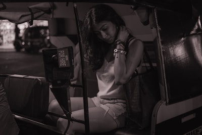A person is seated inside a vehicle, likely a tuk-tuk or auto rickshaw. The individual appears to be in a pensive or contemplative mood, with a visible 'For Hire' sign on the vehicle. The setting seems to be at night or in low light, creating a moody atmosphere.