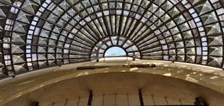 An architectural structure featuring a large, intricate glass and metal dome. The dome has a fan-like pattern with triangular glass panels framed by metal, allowing natural light to illuminate the interior. The view is from below, emphasizing the symmetry and geometric design.