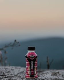 A bottle of BODYARMOR sports drink, labeled strawberry banana, stands on a rocky surface against a blurred backdrop of mountains and a soft, gradient sky.