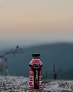 A bottle of BODYARMOR sports drink, labeled strawberry banana, stands on a rocky surface against a blurred backdrop of mountains and a soft, gradient sky.