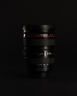 A camera lens placed upright against a dark background. The lens has clear details, including brand markings and specifications such as 24-105mm and an aperture of 1:4. The focus rings and zoom markings are also visible, with a red ring indicating a premium model.