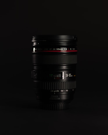 A camera lens placed upright against a dark background. The lens has clear details, including brand markings and specifications such as 24-105mm and an aperture of 1:4. The focus rings and zoom markings are also visible, with a red ring indicating a premium model.