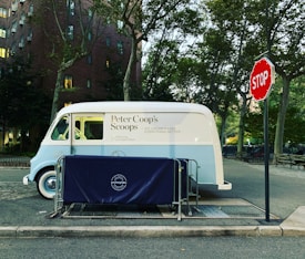 An ice cream truck parked on the side of a street next to a stop sign. The truck is white and light blue, with 'Peter Coop’s Scoops' written on its side. It is surrounded by trees and buildings, and there is a protective blue barrier with 'good neighbors' branding in front of it.