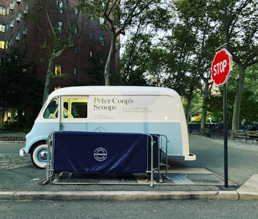 An ice cream truck parked on the side of a street next to a stop sign. The truck is white and light blue, with 'Peter Coop’s Scoops' written on its side. It is surrounded by trees and buildings, and there is a protective blue barrier with 'good neighbors' branding in front of it.
