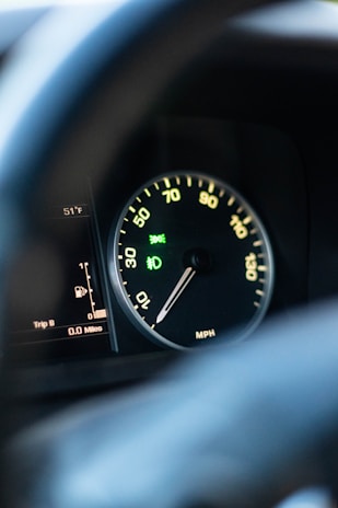 A close-up view of a car speedometer and odometer, displaying the speed in miles per hour and trip details. The temperature gauge shows 51°F. The speedometer needle points to zero, and there are dashboard symbols illuminated in green, indicating various alerts.