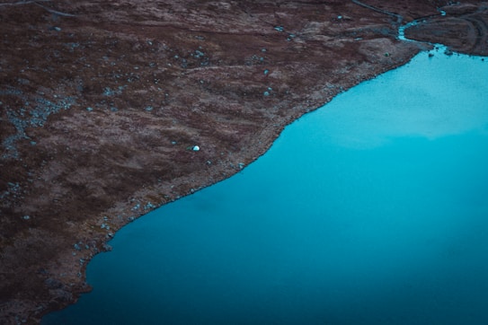 A serene landscape with a large expanse of calm, blue water bordered by rugged, brown terrain. Scattered rocks and subtle textures create a natural, untouched look along the shoreline.
