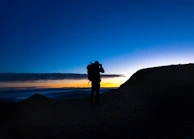 A winding mountain trail at dusk with a lone traveler’s silhouette against a blood-red sky.