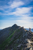 A rugged mountain ridge with steep slopes covered in rocky terrain and patches of green grass. Two hikers are carefully making their way along the ridge, surrounded by a clear blue sky with scattered clouds.
