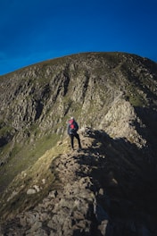 A hiker adjusting their backpack on a rocky mountain trail under a clear blue sky.