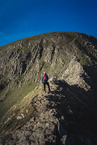 A hiker adjusting their backpack on a rocky mountain trail under a clear blue sky.