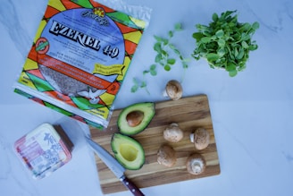 A kitchen scene with various food items including a package of Ezekiel 4:9 tortillas, an avocado cut in half, several mushrooms, a bunch of leafy greens, and a block of tofu. A knife rests on a wooden cutting board beside the avocado and mushrooms.