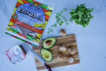 A kitchen scene with various food items including a package of Ezekiel 4:9 tortillas, an avocado cut in half, several mushrooms, a bunch of leafy greens, and a block of tofu. A knife rests on a wooden cutting board beside the avocado and mushrooms.