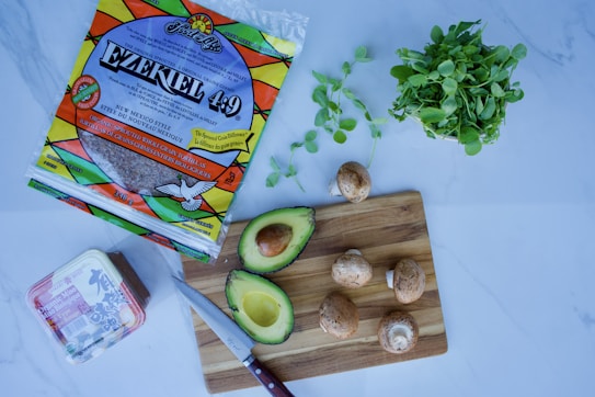 A kitchen scene with various food items including a package of Ezekiel 4:9 tortillas, an avocado cut in half, several mushrooms, a bunch of leafy greens, and a block of tofu. A knife rests on a wooden cutting board beside the avocado and mushrooms.