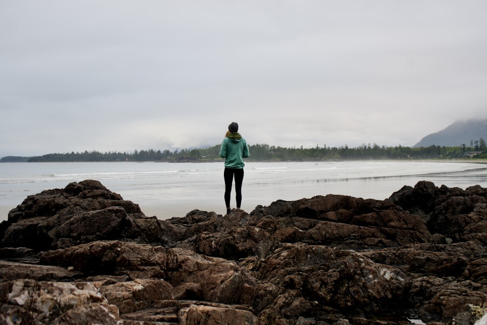 man in green jacket standing on brown rock near body of water during daytime