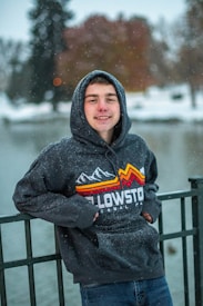A person wearing a hooded sweatshirt with a Yellowstone National Park design stands outdoors near a snowy water body. Snowflakes are visible, and the background features blurred trees and a fence.