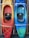 Two kayaks are mounted vertically on a wooden rack. The kayak on the left has a gradient from yellow to orange, while the one on the right transitions from blue to green. Both kayaks are equipped with cushioned seats and elastic straps for securing gear.